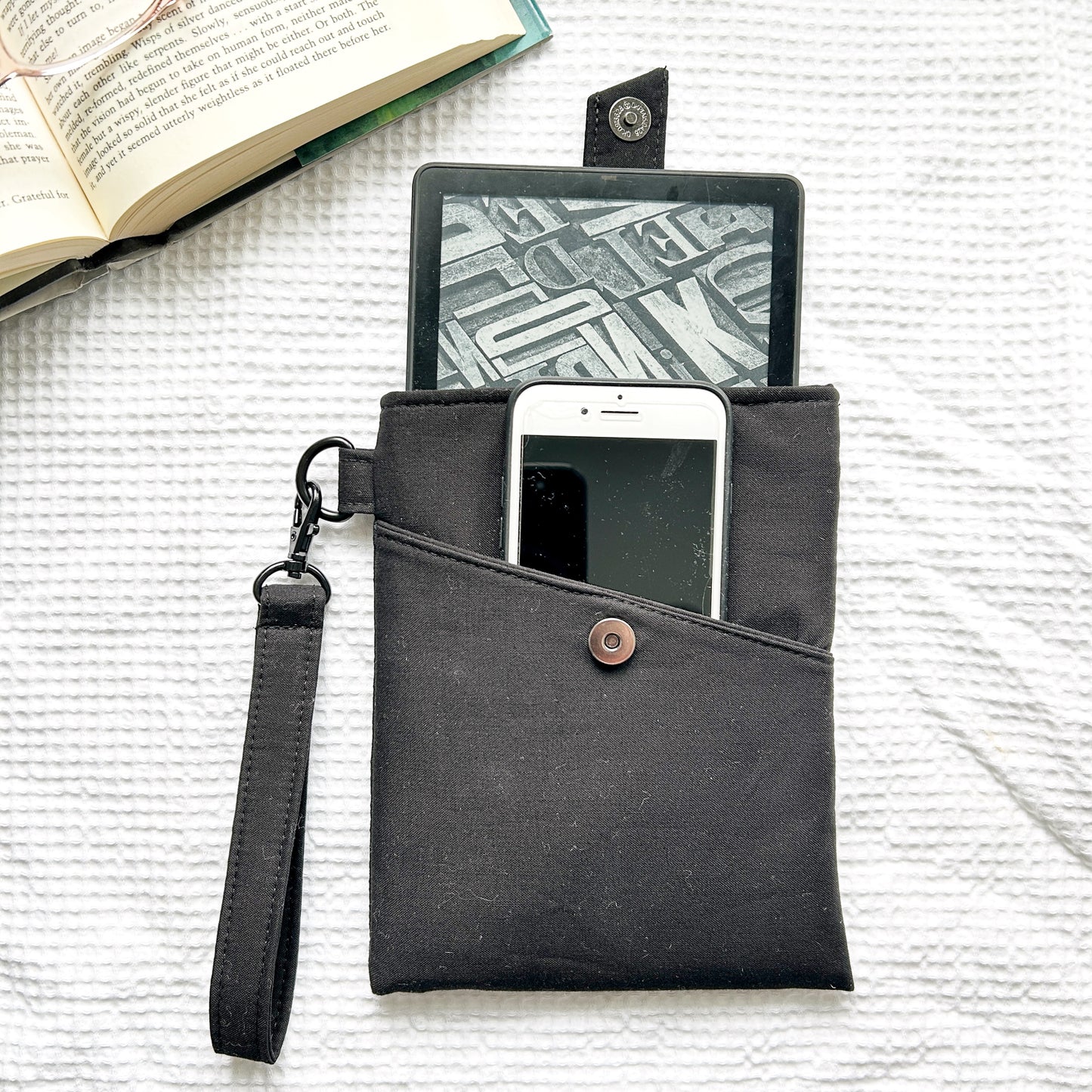 Black phone pouch with a strap, containing a phone and tablet, on a textured surface with an open book.